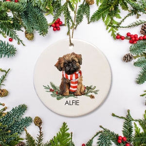 Round ceramic ornament with a Brussels Griffon dog illustration wearing a scarf, surrounded by Christmas foliage. Personalized with your pet's name
